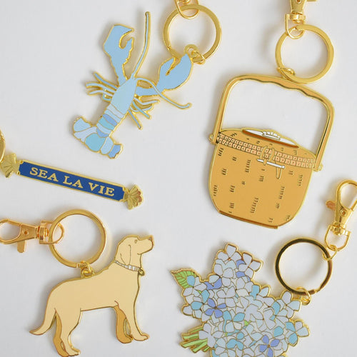Six imported Sara Fitz gold keychains—a Blue Lobster Bag Charm, a woven basket, a “Sea La Vie” charm, a yellow dog, a bouquet of blue flowers, and a ring clasp—feature vibrant enamel details and are arranged on a white background.