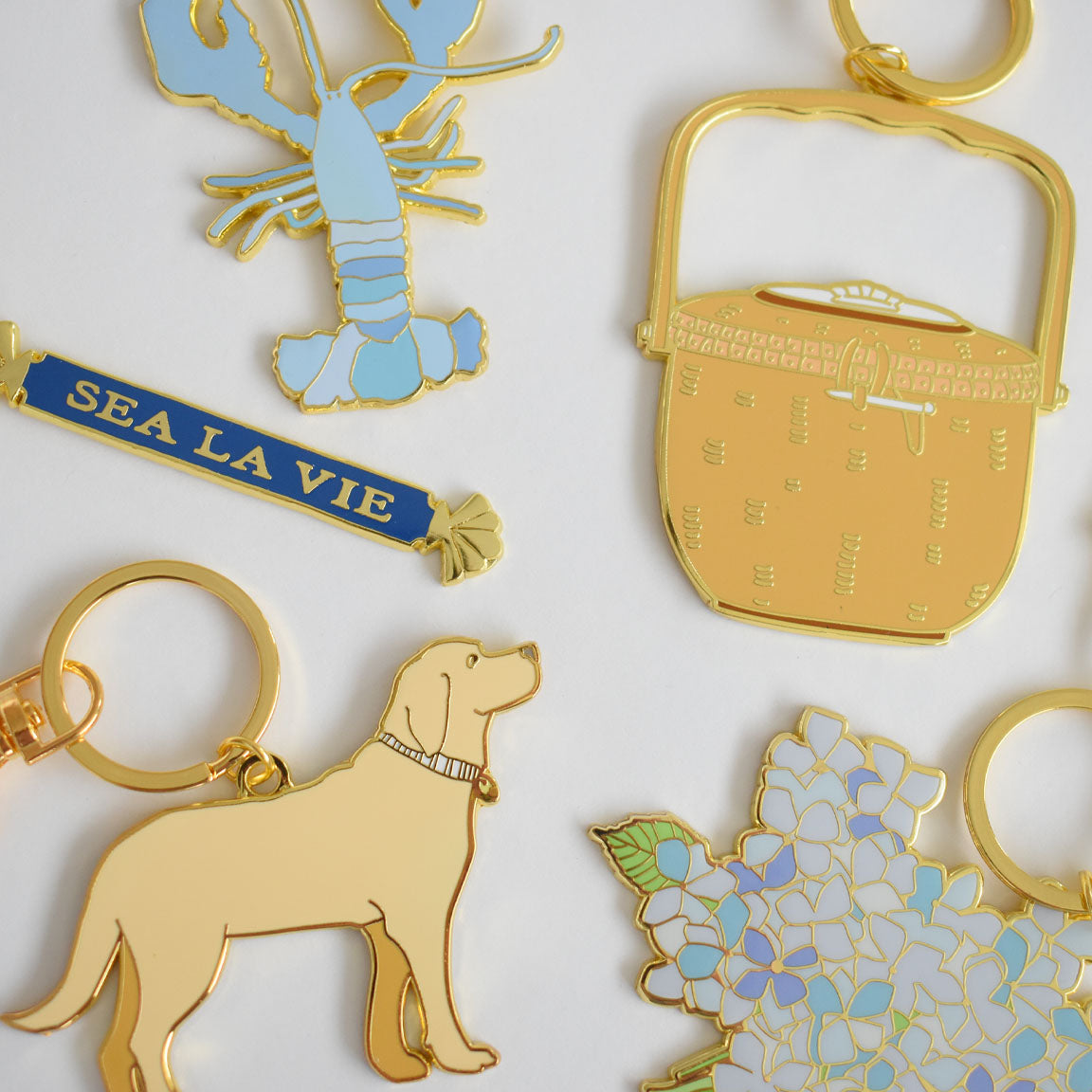 Five imported gold Quarterboard Bag Charms by Sara Fitz are displayed on a white background, featuring enamel designs of a blue lobster, a picnic basket, a SEA LA VIE banner, a yellow dog, and blue hydrangea flowers.