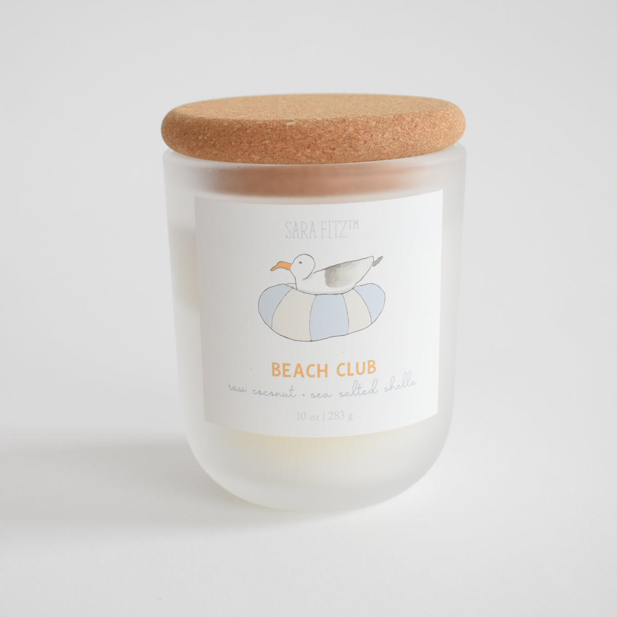 The Sara Fitz Beach Club Candle is a 10 oz frosted glass coconut soy wax candle with a cork lid, featuring a seagull on a striped float label. Scented with sea coconut & sea salted shells. Hand-poured in Maine for that beachy aroma.