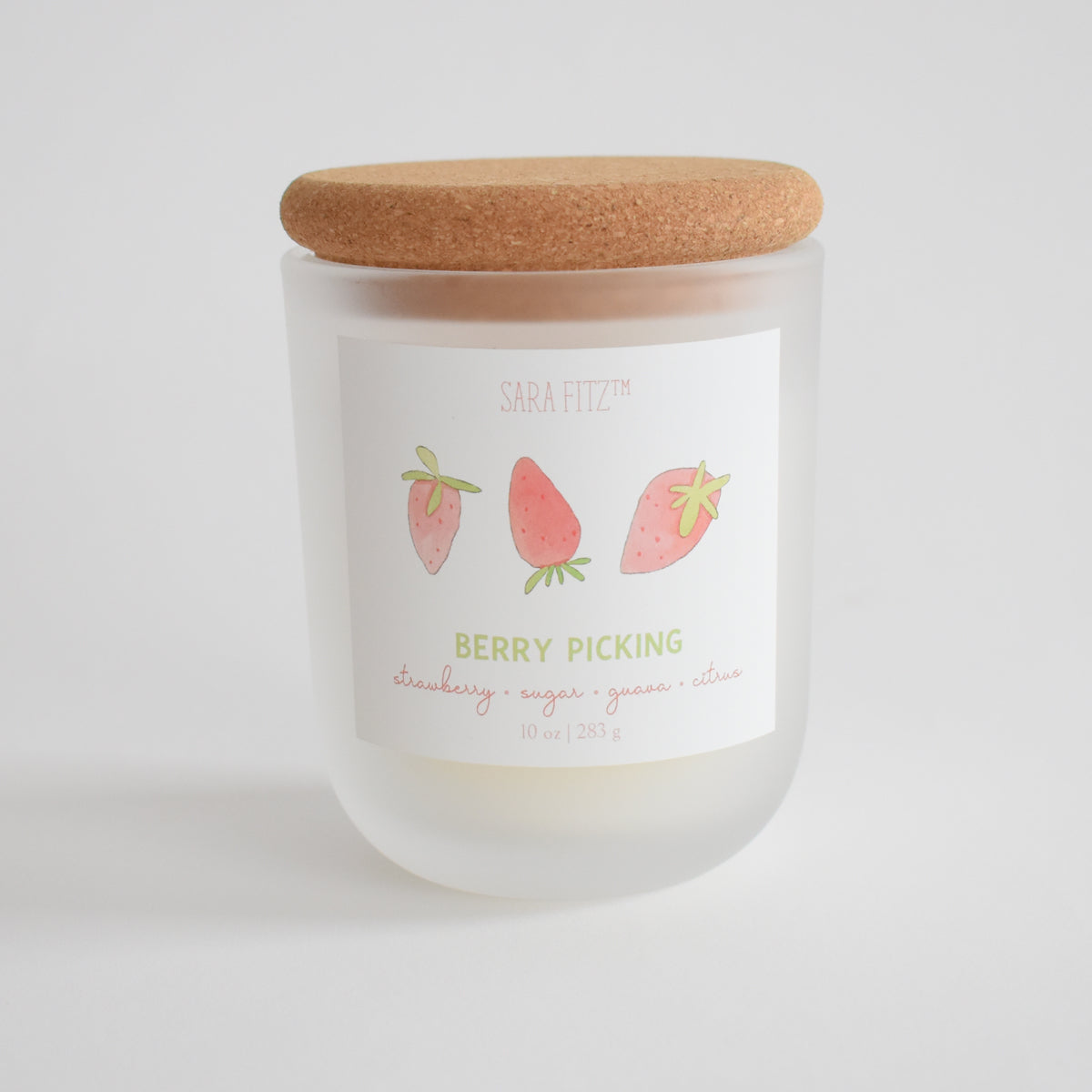 The Berry Picking Candle by Sara Fitz is a 10 oz frosted glass jar with a cork lid, featuring hand-drawn strawberries. Hand-poured in Maine with coconut soy wax, it has notes of strawberry, sugar, guava, and citrus. Plain white background.