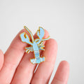 A hand holds the Sara Fitz Blue Lobster Needle Minder, a small enamel pin shaped like a light blue lobster with gold outlines, against a plain white background. This charming accessory is perfect for keeping your needle secure and stylish while you work on your latest stitching project.