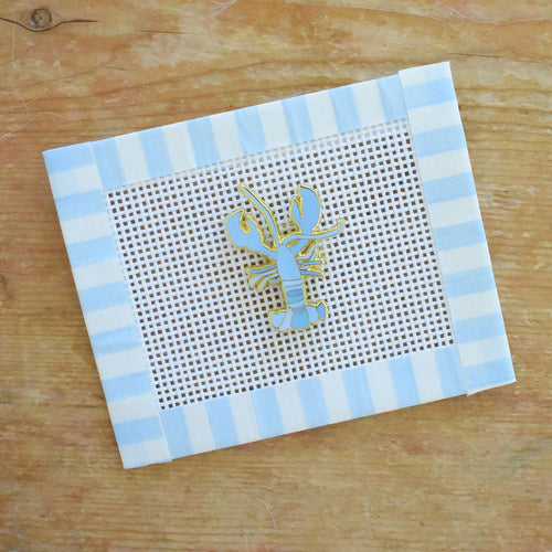 A Blue Lobster Needle Minder by Sara Fitz is placed on a piece of gridded mesh, bordered by blue and white striped tape, atop a wooden surface. This charming accessory keeps your needle secure and organized during your crafting projects.