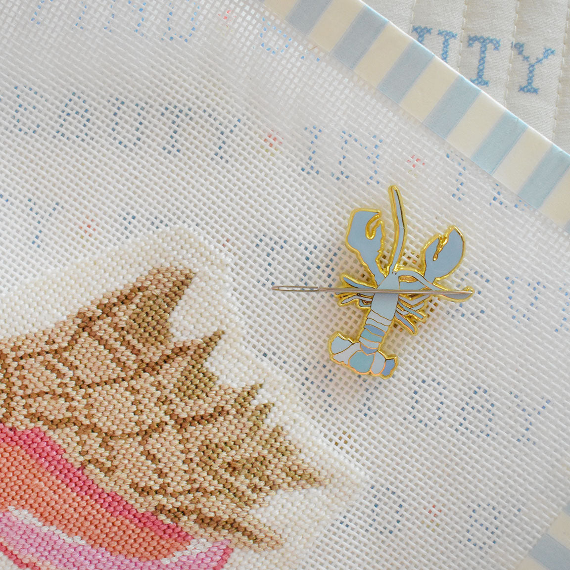 The Blue Lobster Needle Minder by Sara Fitz, a charming pastel blue enamel pin perfect as a stitching accessory or needle holder, is displayed atop a mesh canvas featuring a needlepoint seashell design. Faded text and blue-striped fabric frame the scene, highlighting this delightful enamel needle minder.