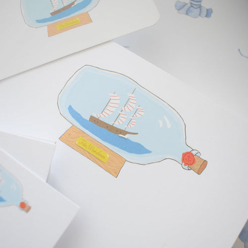 The Boat in a Bottle Notepad by Sara Fitz features an illustration of a sailing ship with red-striped sails in a glass bottle on a wooden stand labeled "The Wanderer," sized 5x7 inches, made in the USA, with other cards partially visible behind.