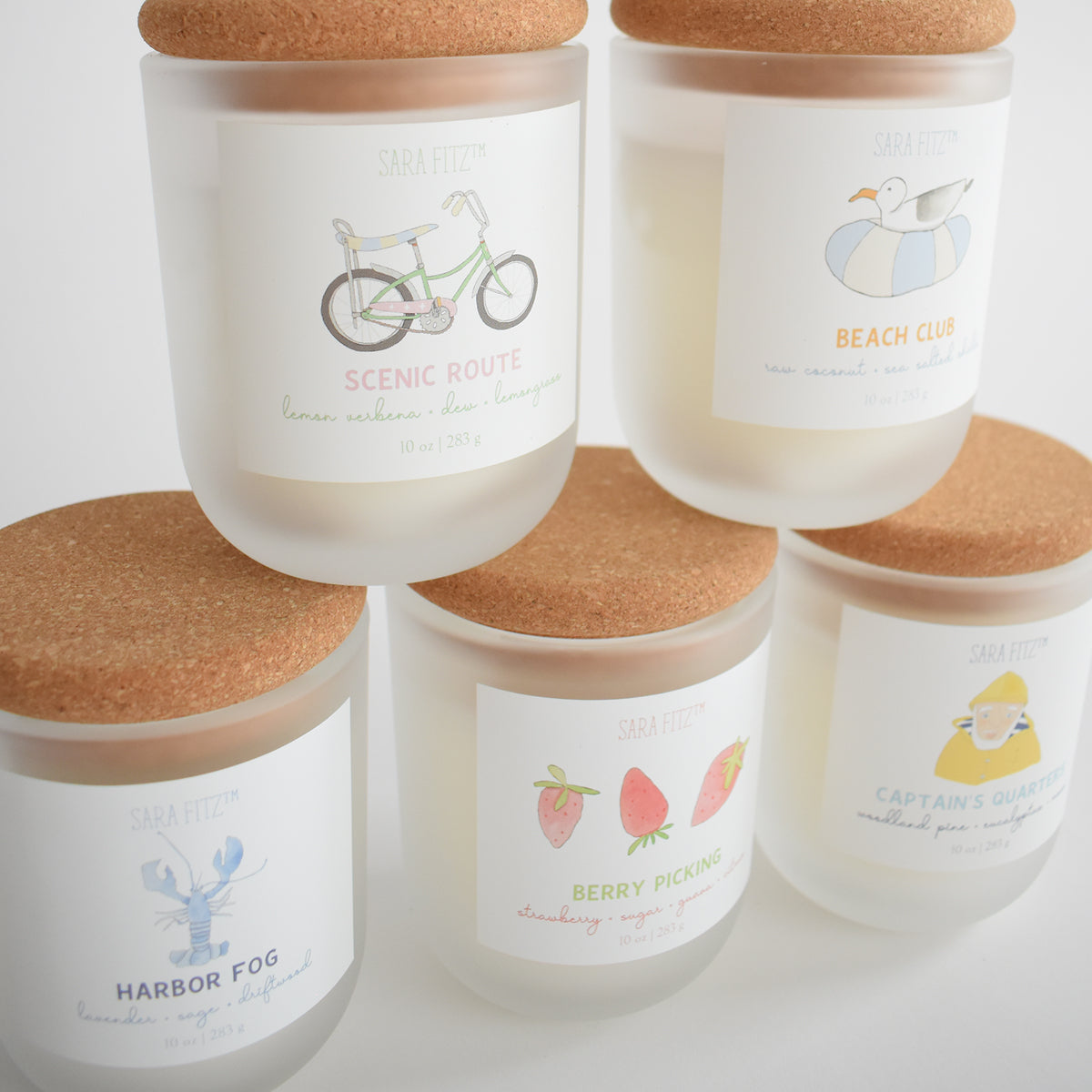Five Captain's Quarters Candles by Sara Fitz, made with coconut soy wax and cork lids, are hand-poured in Maine. Each frosted glass candle features a unique illustrated label: bicycle, bird, lobster, strawberries, or seashells.