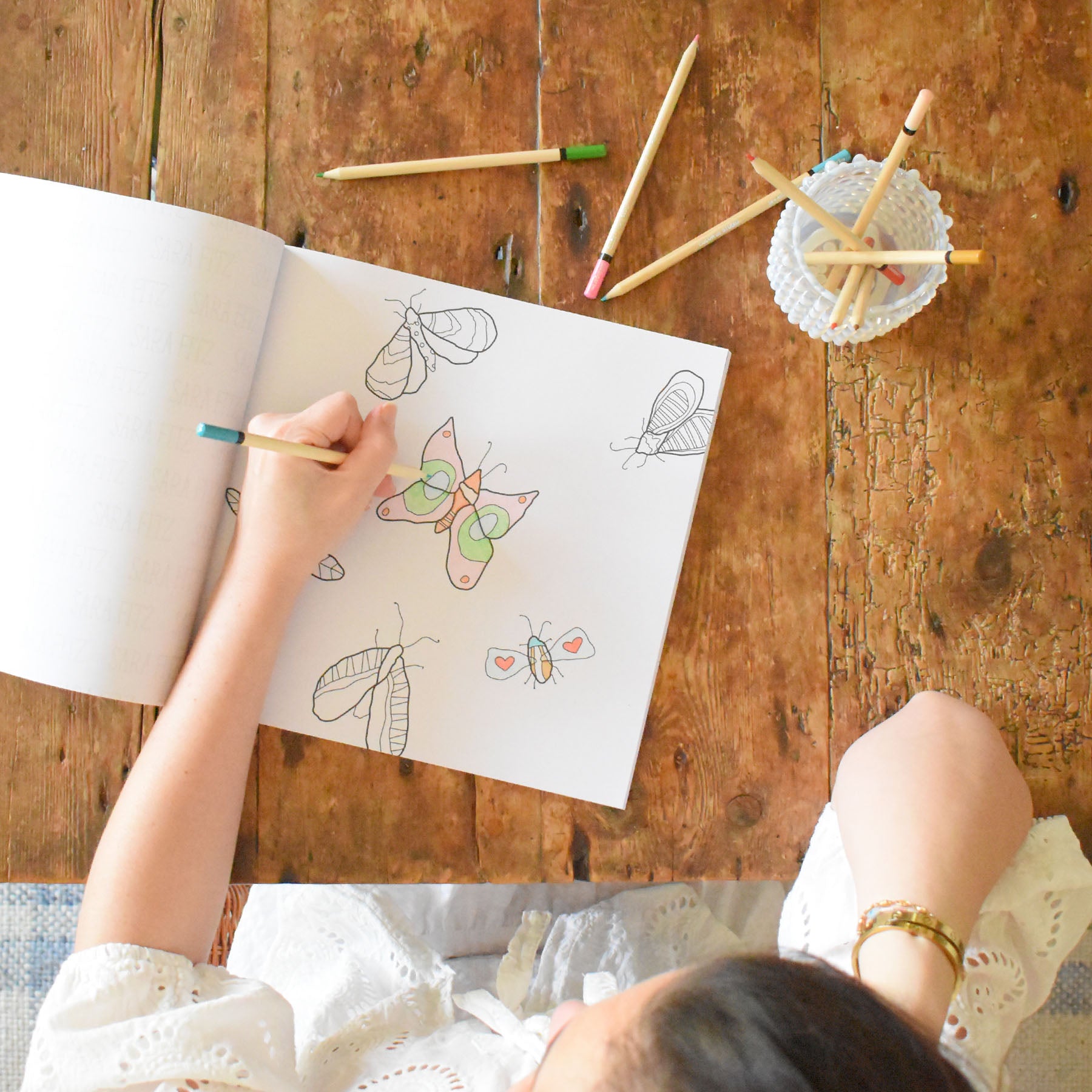 A person coloring butterfly drawings from the Sara Fitz Coloring Book Countryside on a wooden table, with colored pencils and a small bowl of pencils nearby.