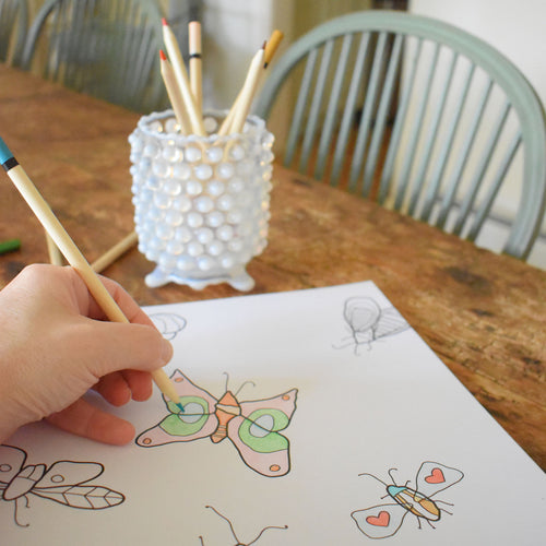 A hand colors a butterfly drawing on white paper at a wooden table, with a white textured cup holding colored pencils and additional butterfly sketches nearby. Reminiscent of classic New England illustrations, the scene evokes the charm of the Sara Fitz “Coloring Book Countryside,” an adult coloring book designed for relaxation. Pale green chairs are in the background.