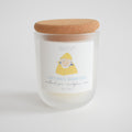 The Sara Fitz Captain's Quarters Candle features a woodland pine, eucalyptus, and moss scent, hand-poured in Maine with coconut soy wax. It comes in a frosted glass jar with cork lid and a label showing a person in a yellow raincoat.