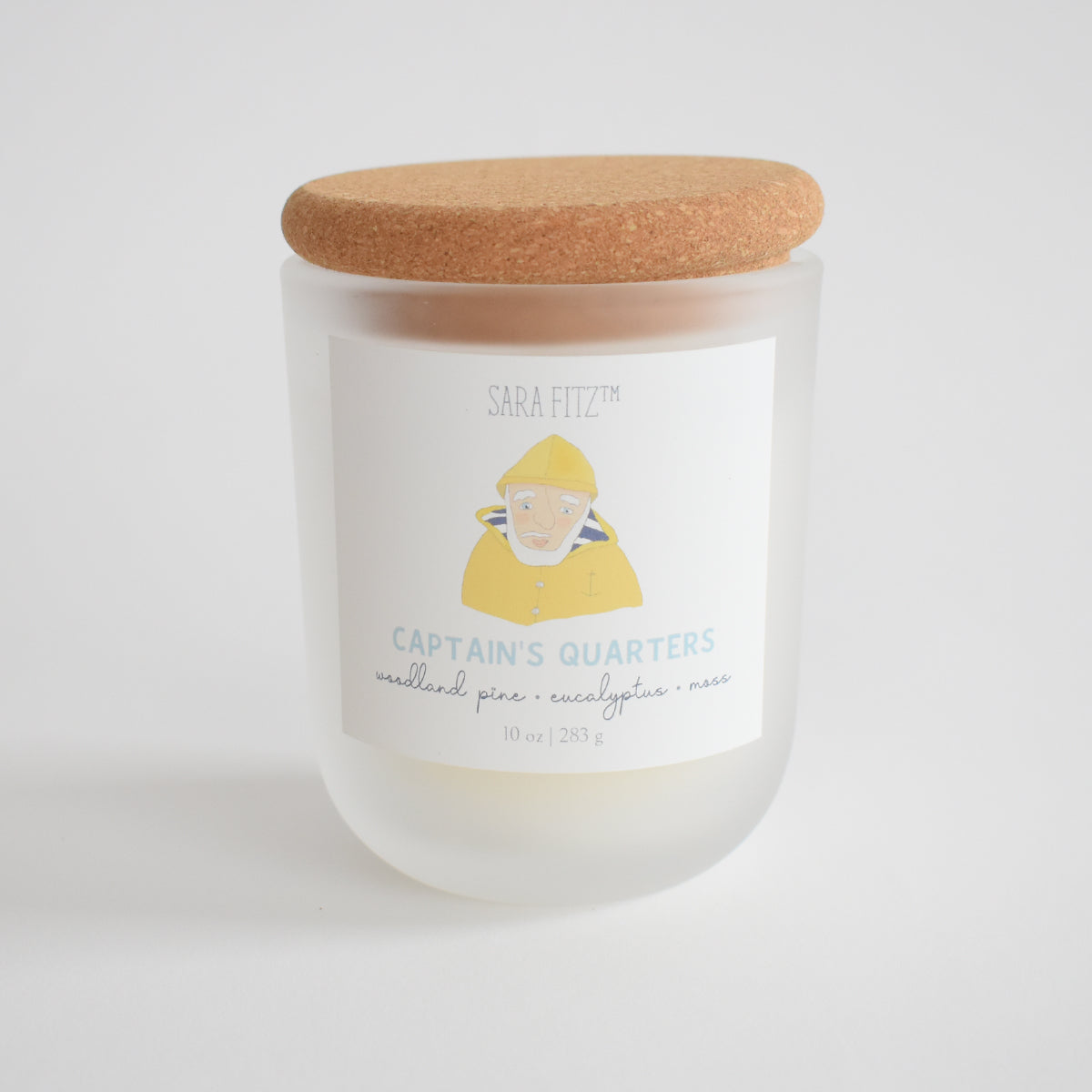 The Sara Fitz Captain's Quarters Candle features a woodland pine, eucalyptus, and moss scent, hand-poured in Maine with coconut soy wax. It comes in a frosted glass jar with cork lid and a label showing a person in a yellow raincoat.