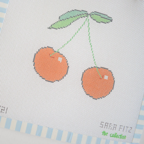 The Cherry Needlepoint by Sara Fitz features two orange cherries with green stems on a white background, bordered by light blue and white stripes. "Sara Fitz" and the collection are printed in the lower right on 18 mesh canvas.