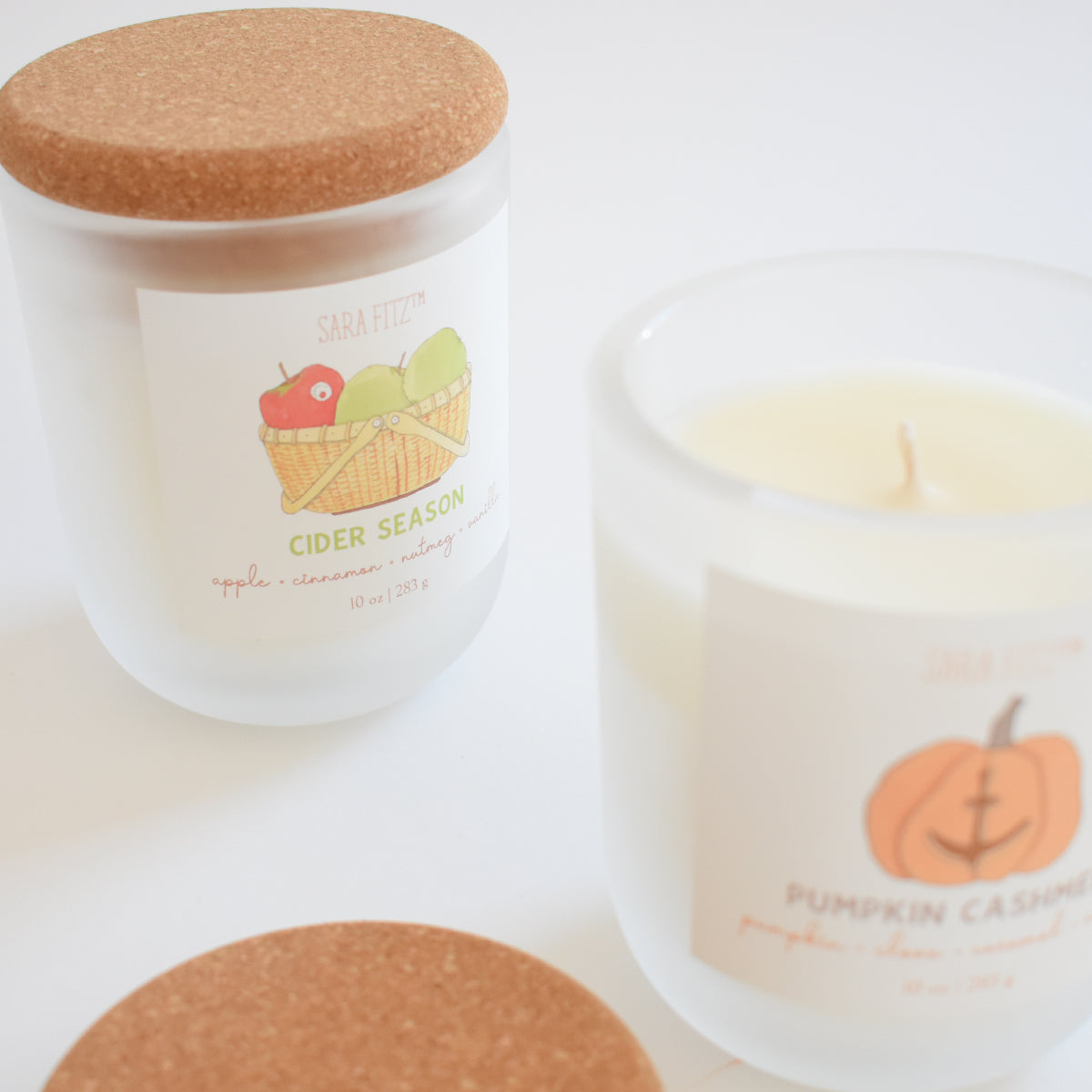 Elevate your space with two white candles in frosted glass jars topped with cork lids from Sara Fitz. The Cider Season Candle displays a label of apples nestled in a basket, hand-poured in Maine and scented with apple cinnamon for a warm seasonal aroma. Its companion features an adorable pumpkin illustration and the Pumpkin Cashew scent. Both are crafted from premium coconut soy wax to ensure a clean, inviting burn.