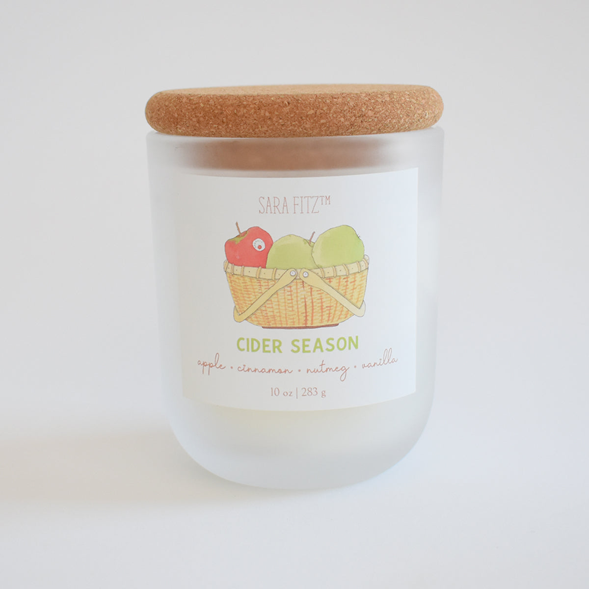 Hand-poured in Maine, the Sara Fitz Cider Season Candle comes in a frosted glass jar with a cork lid. The label reads “SARA FITZ CIDER SEASON” and displays an illustration of a basket holding an apple and a pear. Crafted from coconut soy wax, this apple cinnamon candle features scent notes of apple, cinnamon, nutmeg, and vanilla.