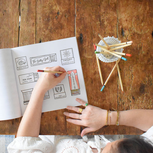 A person colors a page in the Sara Fitz Coloring Book Seaside on a rustic wooden table, with colored pencils in a cup nearby. The pages feature charming New England illustrations that inspire creativity and relaxation.