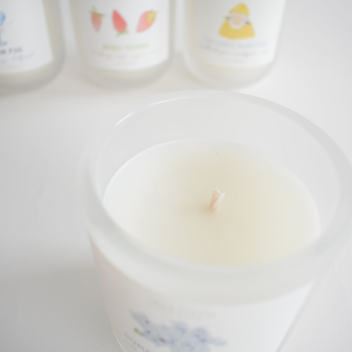 A close-up of the Sara Fitz Harbor Fog Candle, a white coconut soy wax candle in a frosted glass container, hand-poured in Maine. Three other Sara Fitz candles with vibrant fruit or floral label illustrations appear slightly blurred in the background.