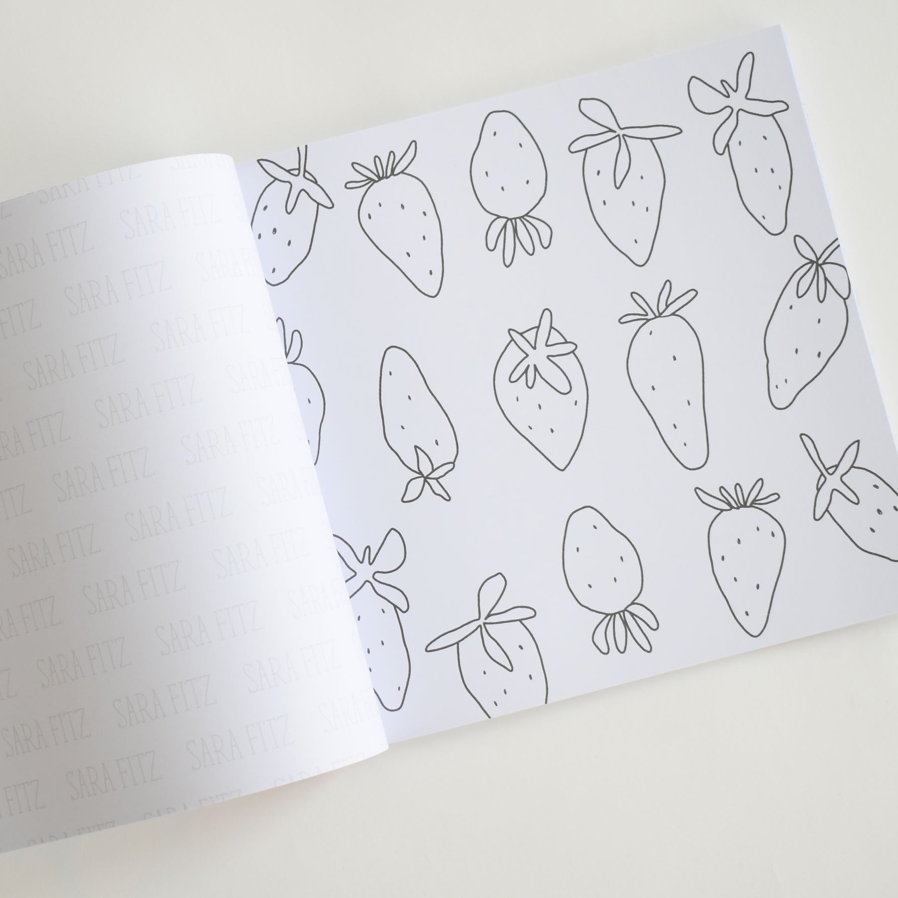 An open Coloring Book Countryside by Sara Fitz displays pages featuring outlined strawberries, ready to be colored. The left page is partially turned, with a subtle SARA FITZ watermark pattern repeated across it. Set against a light surface, the scene captures the charm of New England illustrations—ideal for adult coloring book enthusiasts.