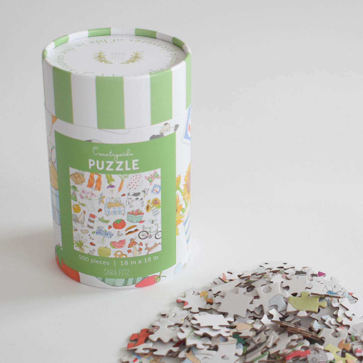 A cylindrical Countryside Puzzle box with a green and white striped lid rests beside a vintage collage of 500 loose jigsaw pieces.