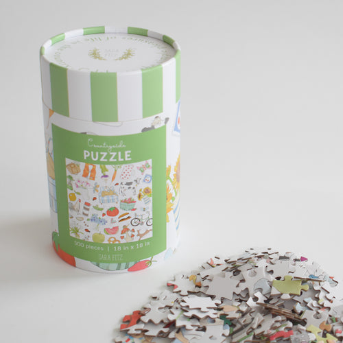 A cylindrical Countryside Puzzle box with a green and white striped lid rests beside a vintage collage of 500 loose jigsaw pieces.