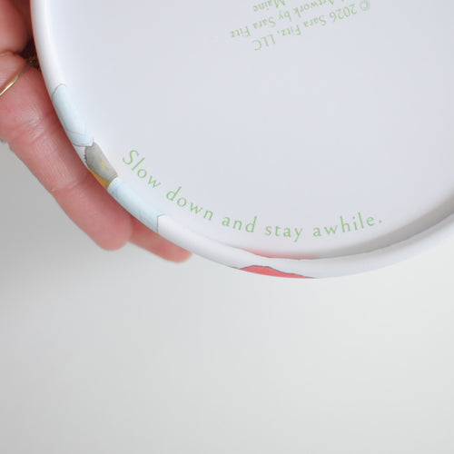 A person holds the round vintage-style lid of a Countryside Puzzle container, printed with the text Slow down and stay awhile.