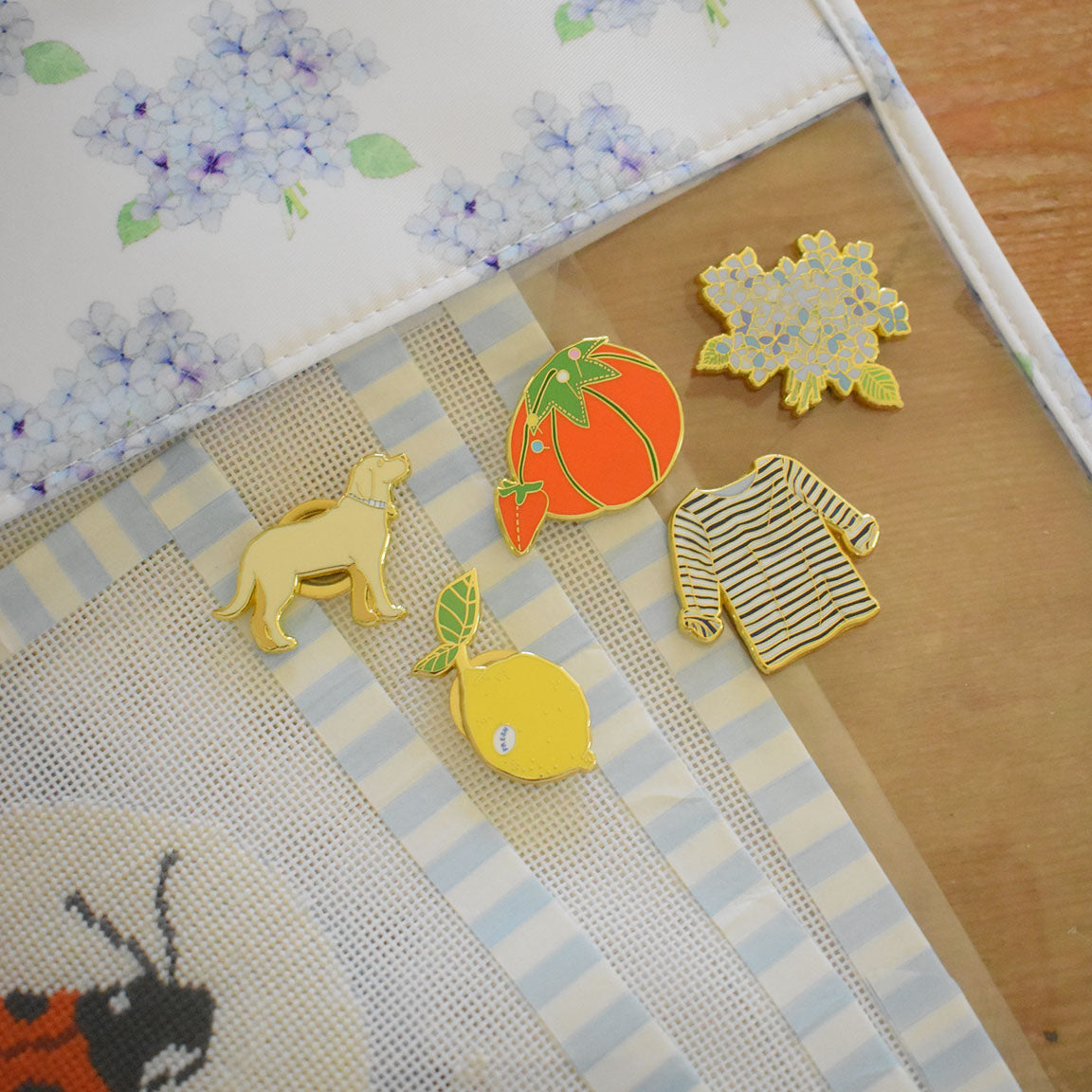 Five enamel pins by Sara Fitz rest on a striped fabric, perfect as stitching accessories. Designs include a dog, a lemon, the Tomato Pincushion Needle Minder, a striped sweater, and a cluster of blue flowers. A floral fabric and part of a ladybug motif are also visible—ideal for anyone seeking unique enamel needle minders or an eye-catching needle holder.