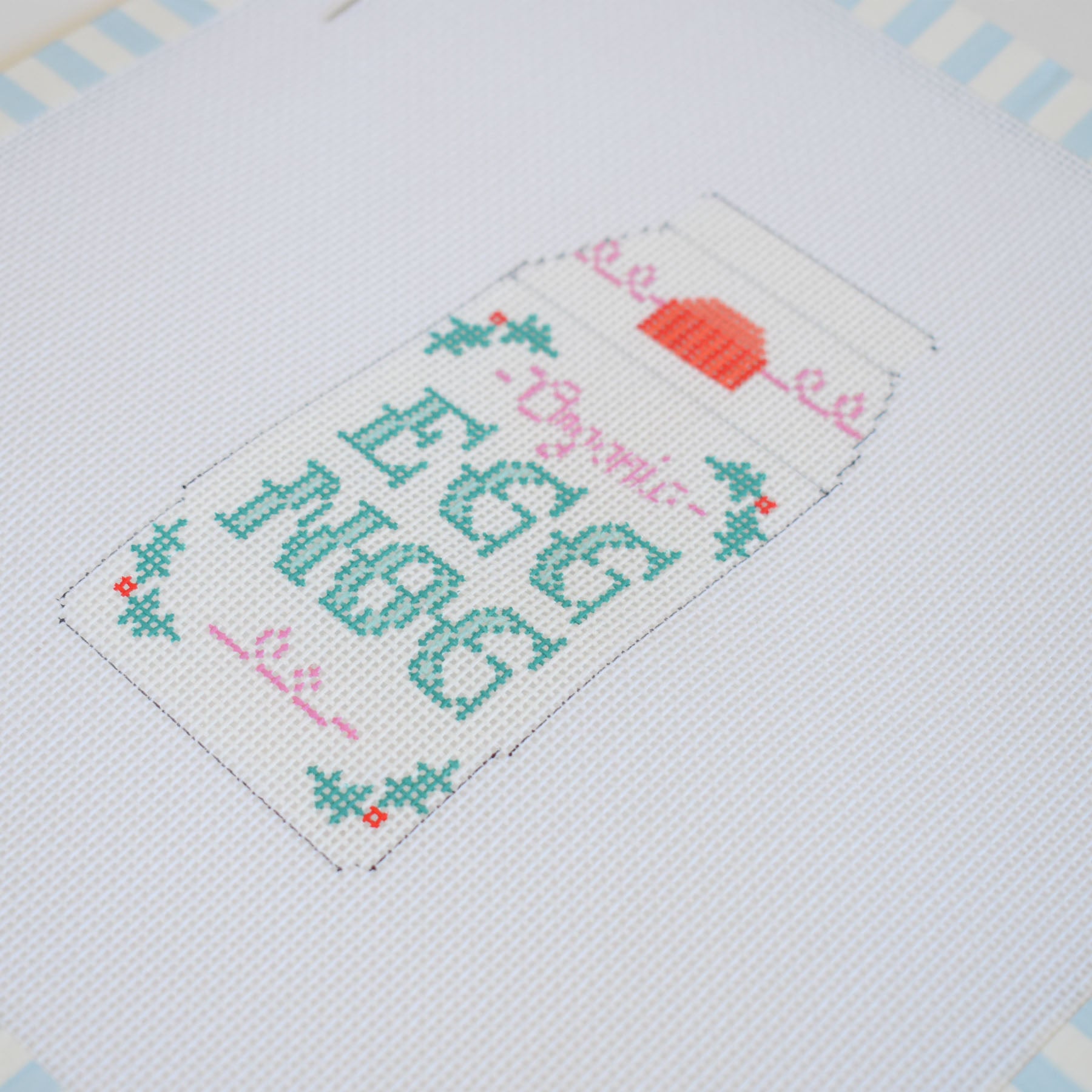 The Eggnog Needlepoint by Sara Fitz features a carton labeled Organic Egg Nog in green and pink, with holly leaves and berries, on white fabric with a blue striped border—ideal for stitching on 18 mesh needlepoint canvas.