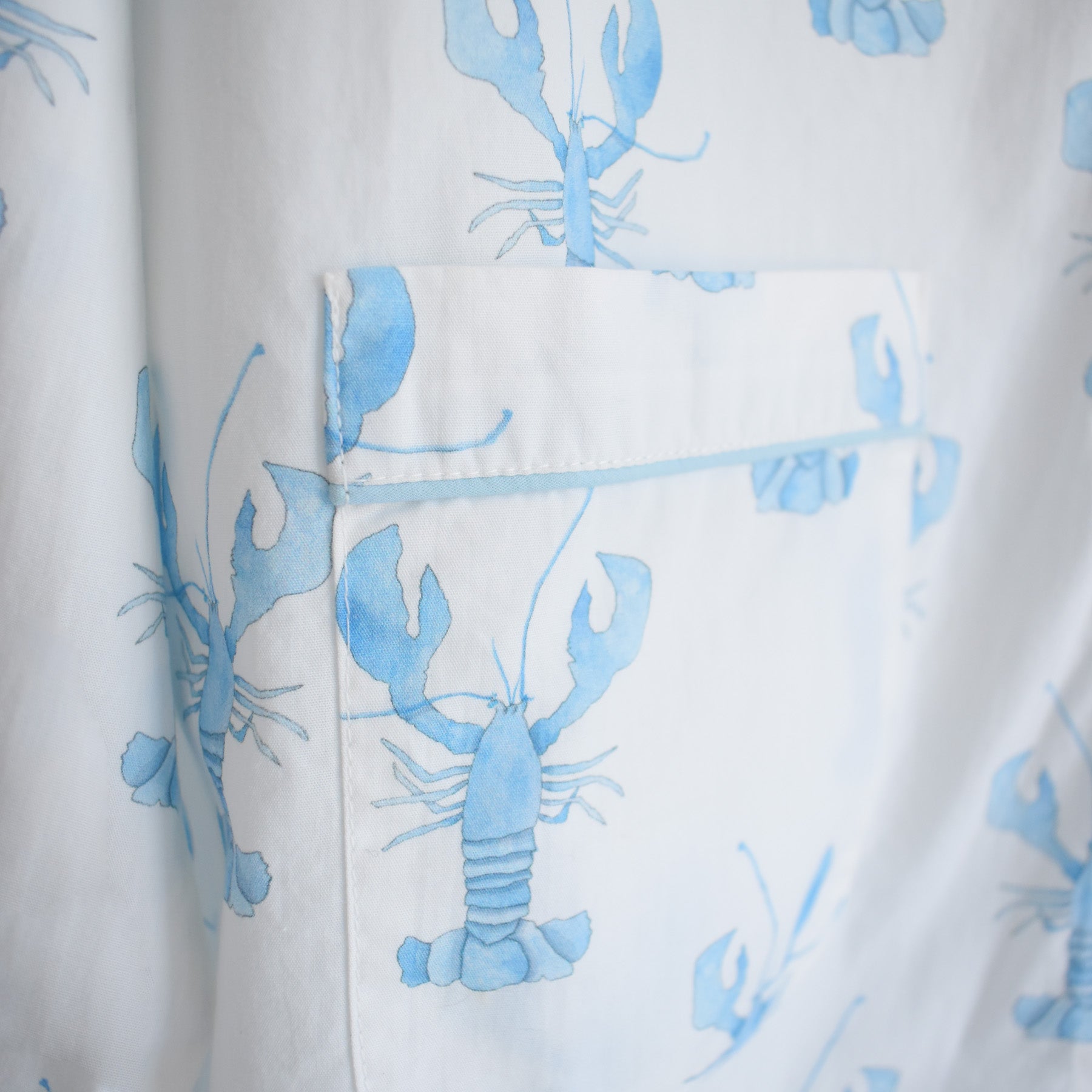 Close-up of a white cotton shirt with a pocket, featuring light blue lobster prints scattered across the fabric, part of the Sara Fitz Blue Lobster Short Pajama Set. This set combines soft comfort with playful coastal style to refresh your sleepwear collection.