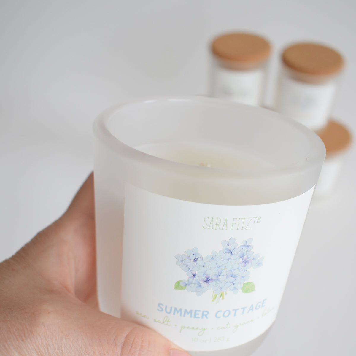 A hand holds a frosted glass coconut soy wax candle labeled "Beach Club" by Sara Fitz, featuring a watercolor design. Three similar, hand-poured-in-Maine candles with wooden lids are blurred in the background.
