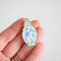 A hand holds the Sara Fitz Ginger Jar Needle Minder, a small enamel pin shaped like a vase and adorned with blue illustrations of a lobster and sea creatures on a white background—perfect as an enamel needle minder or stitching accessory for your needlework projects.