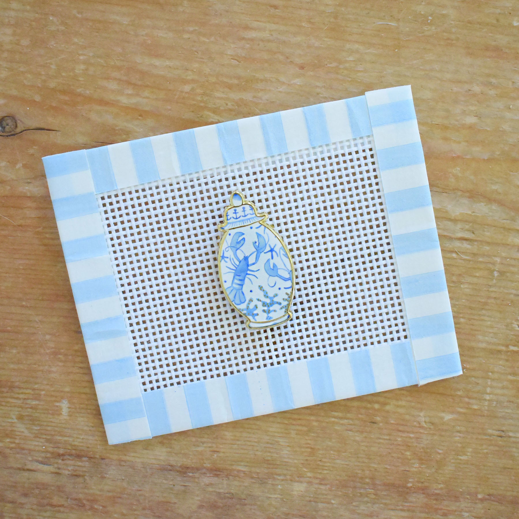 The Ginger Jar Needle Minder by Sara Fitz, featuring a small jar-shaped design in blue and white enamel, rests on a piece of needlepoint canvas bordered with blue and white striped tape atop a wooden surface.