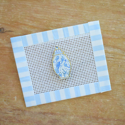The Ginger Jar Needle Minder by Sara Fitz, featuring a small jar-shaped design in blue and white enamel, rests on a piece of needlepoint canvas bordered with blue and white striped tape atop a wooden surface.