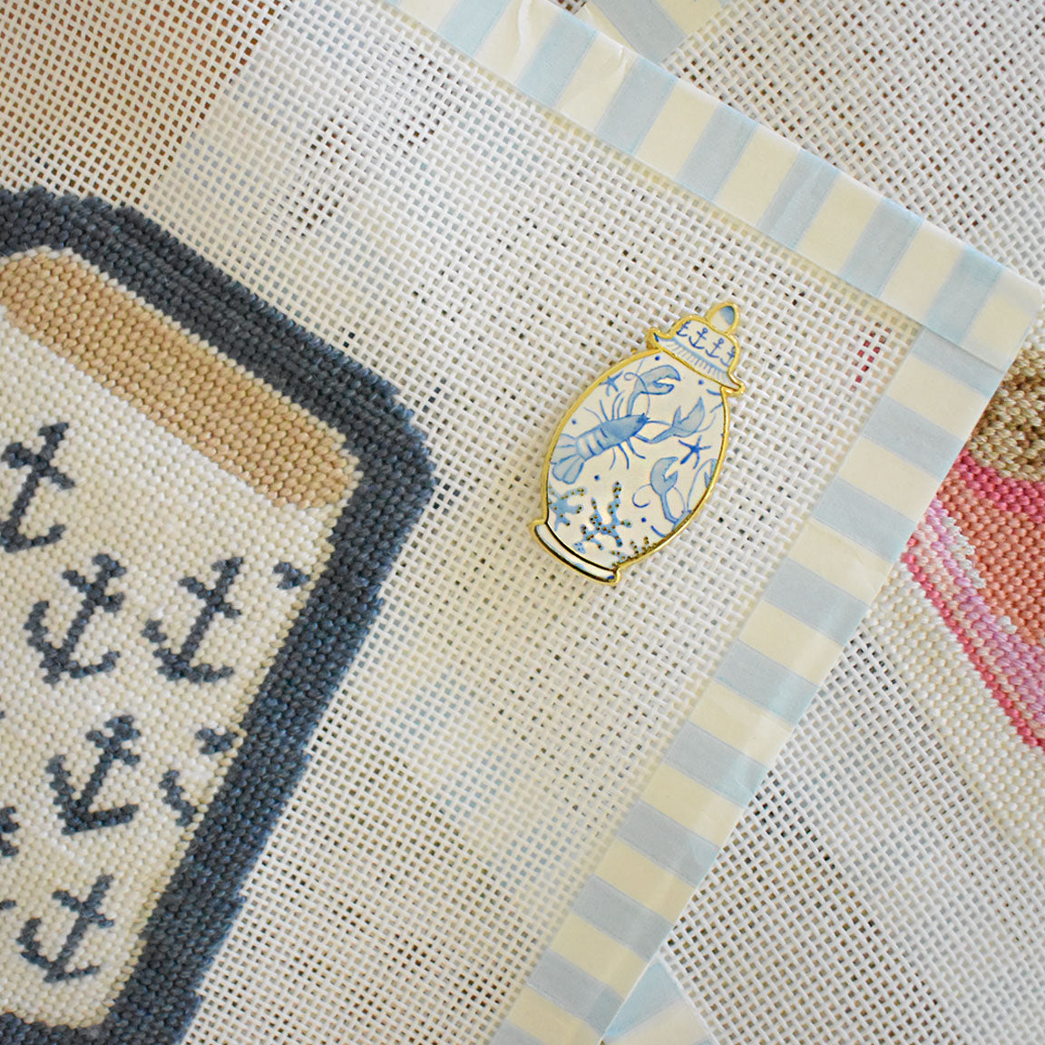 A blue and white Ginger Jar Needle Minder by Sara Fitz rests on mesh fabric adorned with nautical motifs, including a book featuring navy anchors. Perfect for keeping your needle secure while you stitch, this charming enamel accessory combines both style and function. The background showcases light blue and white striped borders.