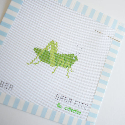 The MINI Grasshopper Needlepoint by Sara Fitz features a stitch-painted green grasshopper on white fabric with light blue striped edges. "SARA FITZ the collection" is printed below. Includes 18 mesh canvas only and a needle at the top right.