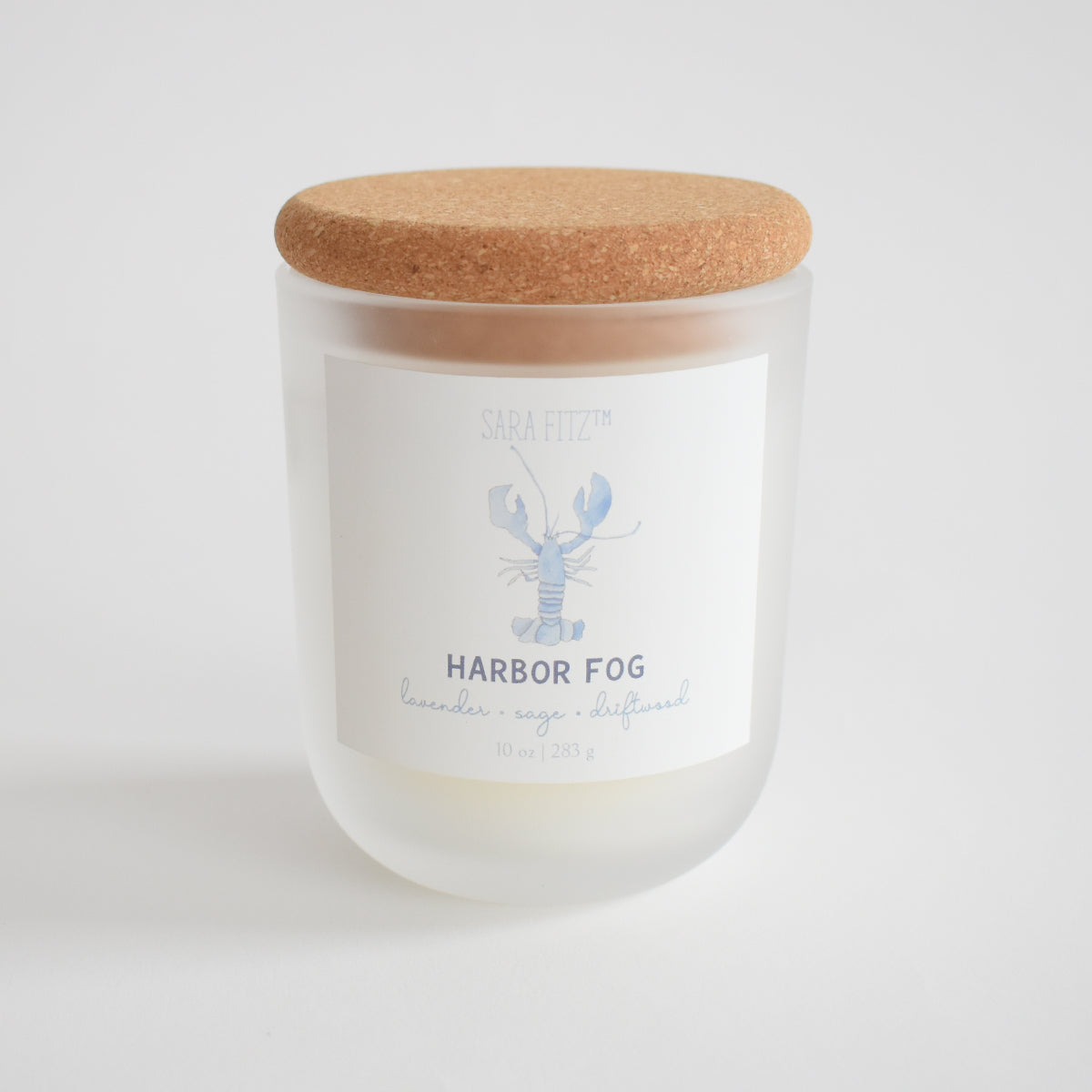The Harbor Fog Candle by Sara Fitz features a frosted glass jar with a cork lid, a light blue lobster label, and scents of lavender, sage, and driftwood. Hand-poured in Maine with coconut soy wax.