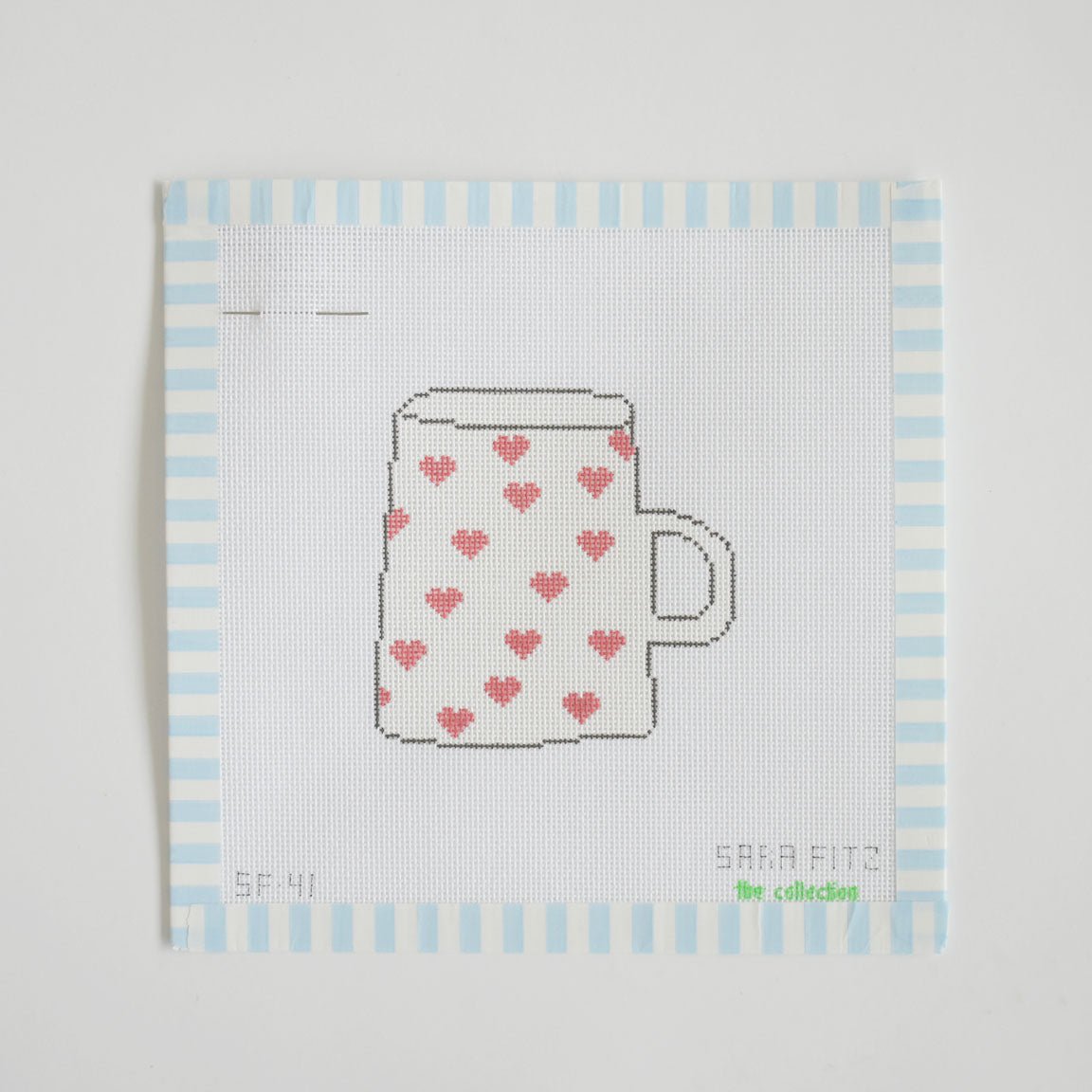 The Heart Mug Needlepoint by Sara Fitz is a stitch-painted cross-stitch pattern featuring a white coffee mug with a handle, adorned with small red hearts, on an 18 mesh canvas square bordered by light blue and white stripes.
