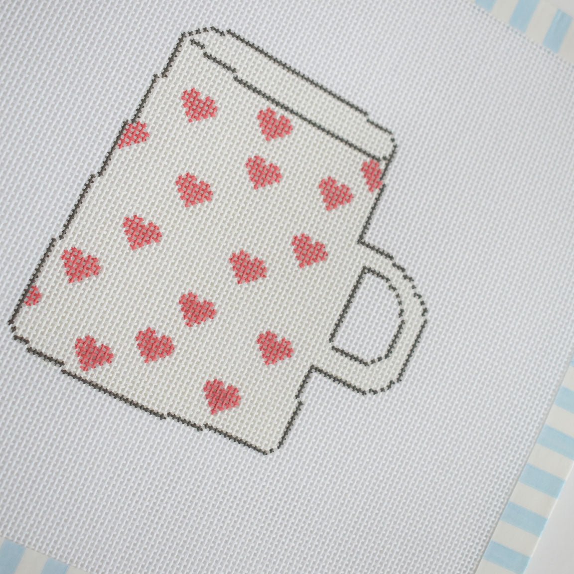 The Heart Mug Needlepoint by Sara Fitz features a cross-stitched image of a white mug adorned with small red hearts, outlined in black, set against white fabric and framed by a blue and white striped border on stitch-painted 18 mesh canvas—ideal for needlepoint enthusiasts.
