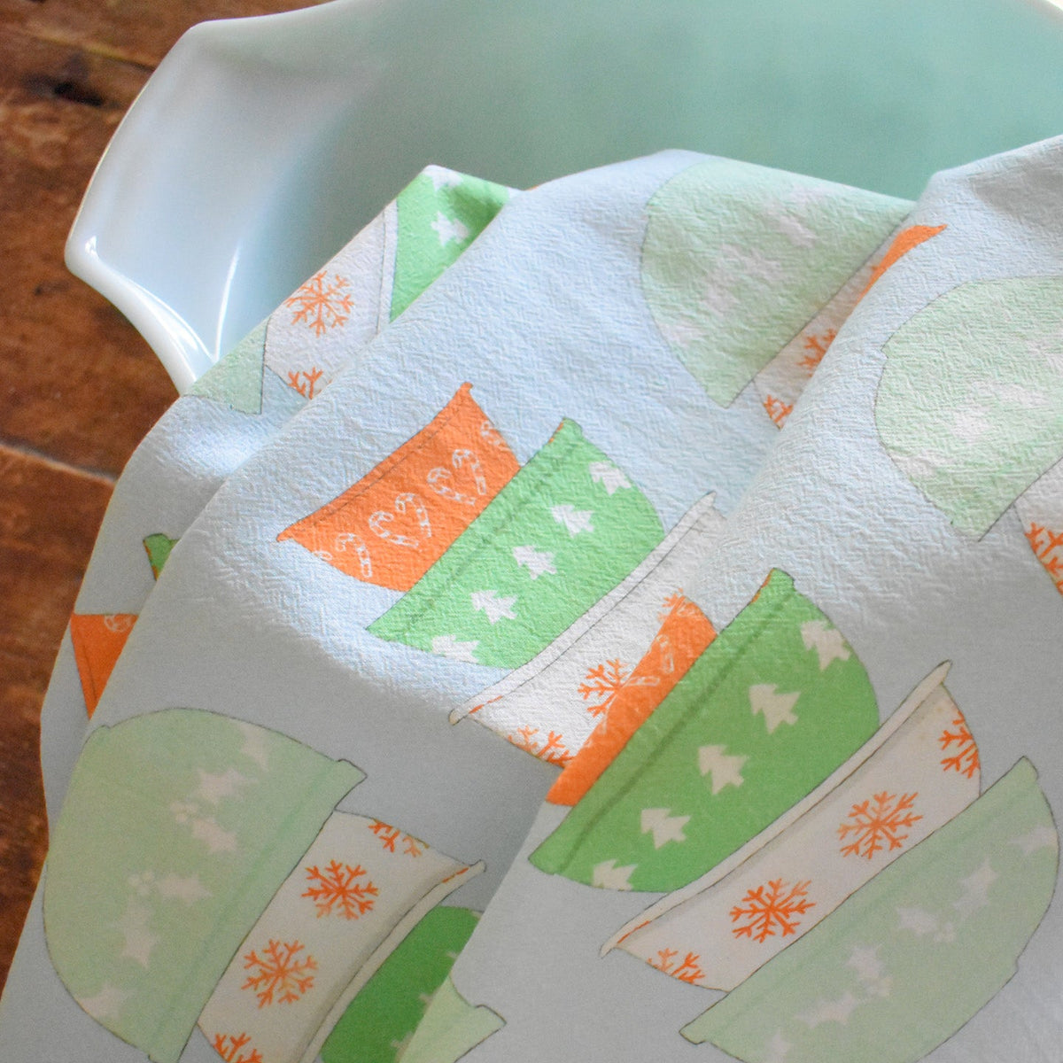 A light blue Heritage Towel, Mixing Bowls organic cotton kitchen towel with a pattern of stacked vintage mixing bowls and snowflakes draped over a ceramic bowl.