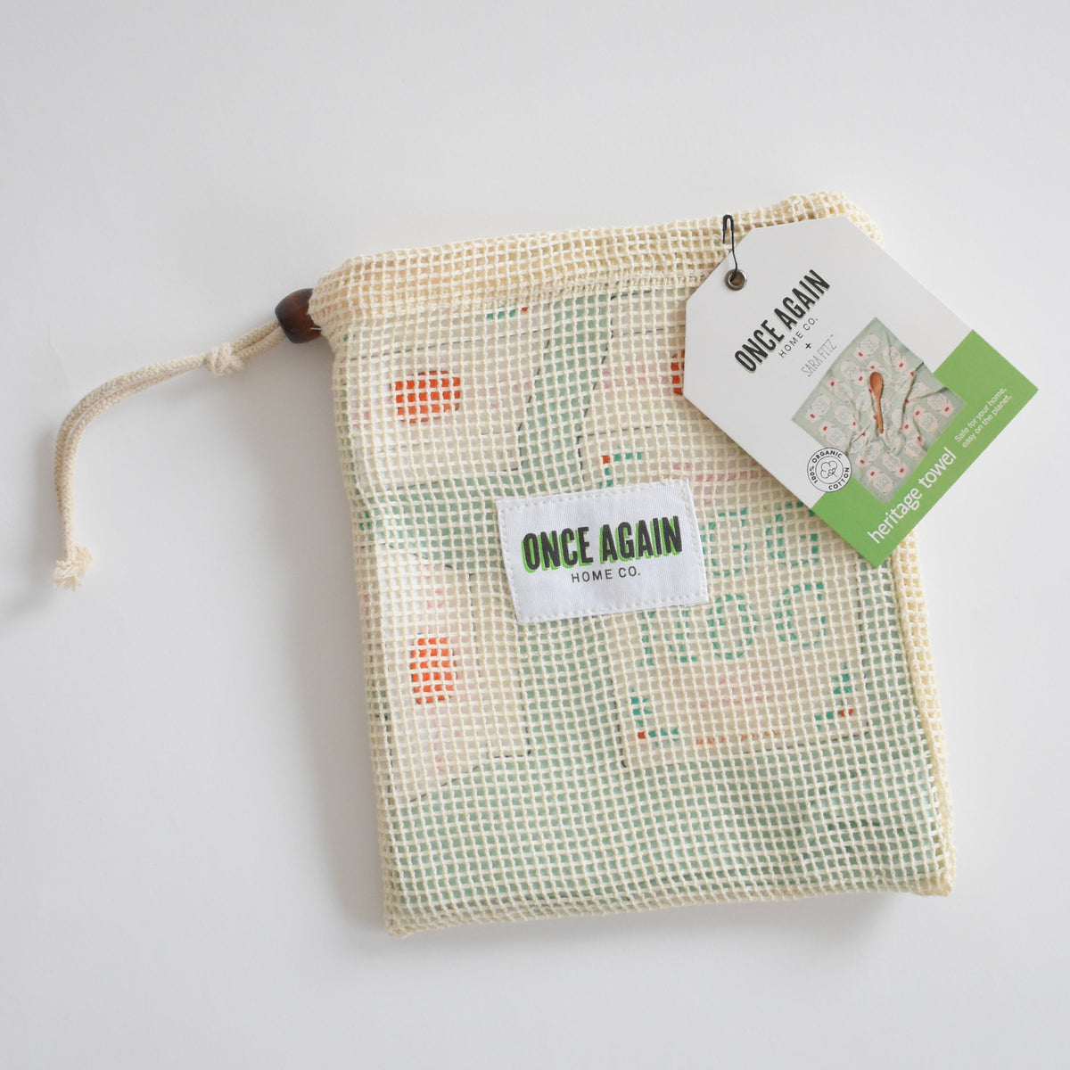 A folded Sara Fitz Heritage Towel in Egg Nog inside a reusable drawstring mesh bag with a brand tag attached.
