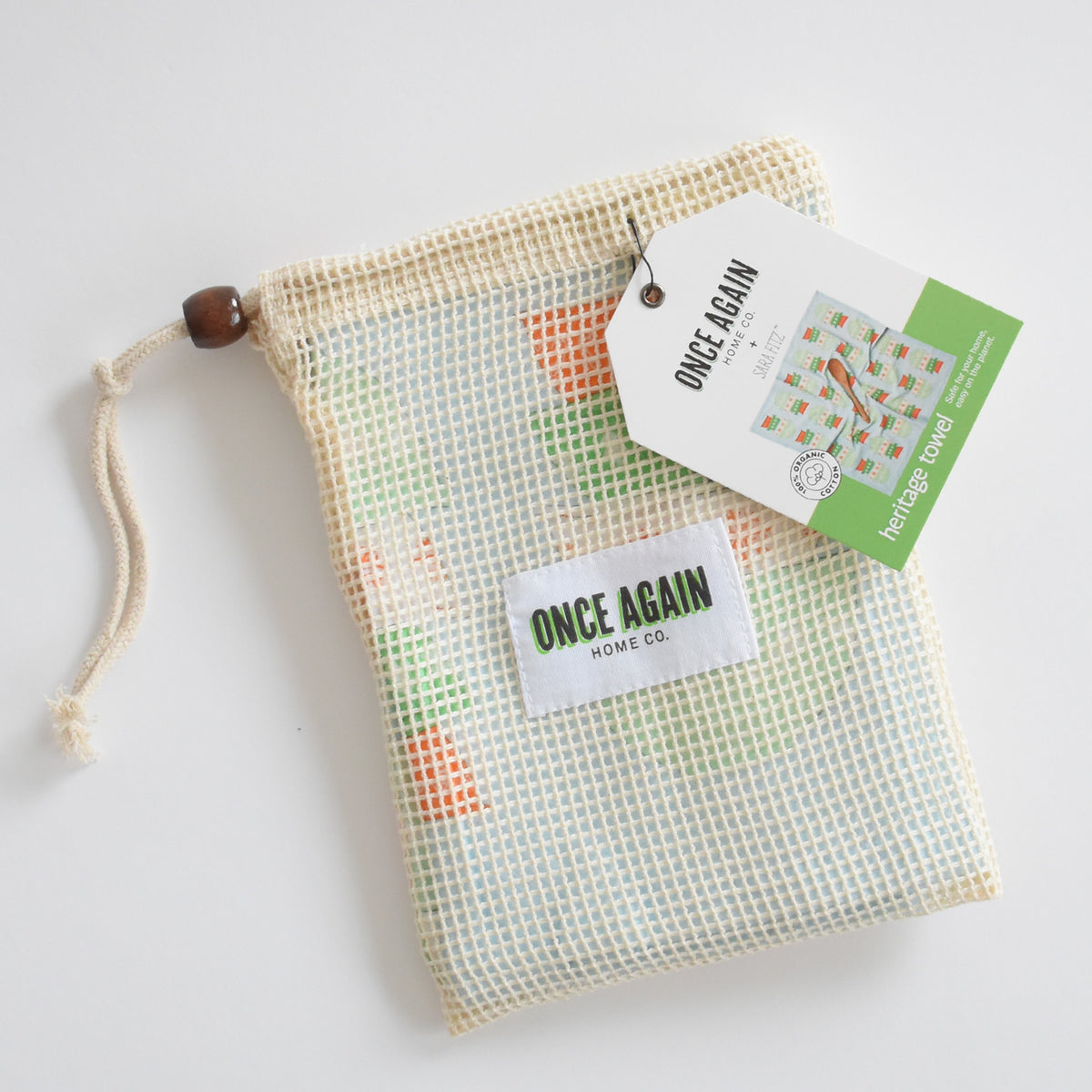 A reusable mesh produce bag containing a Sara Fitz Heritage Towel, with brand tags attached.