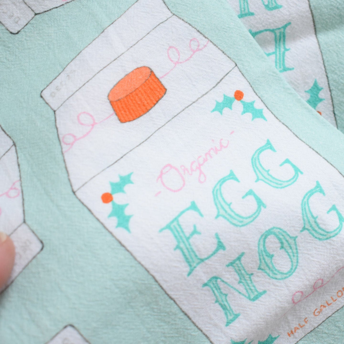 Sara Fitz Heritage Towel, Egg Nog, a light green organic cotton kitchen towel featuring a repeating illustration of an eggnog carton with the word EGG NOG and holly accents.