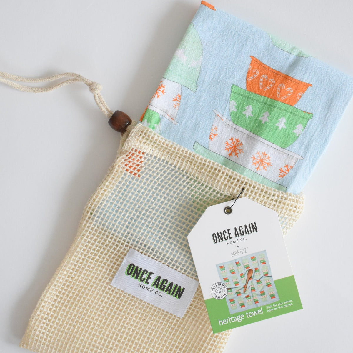 A folded Heritage Towel, Mixing Bowls organic cotton kitchen towel inside a drawstring mesh produce bag with a branded product tag.