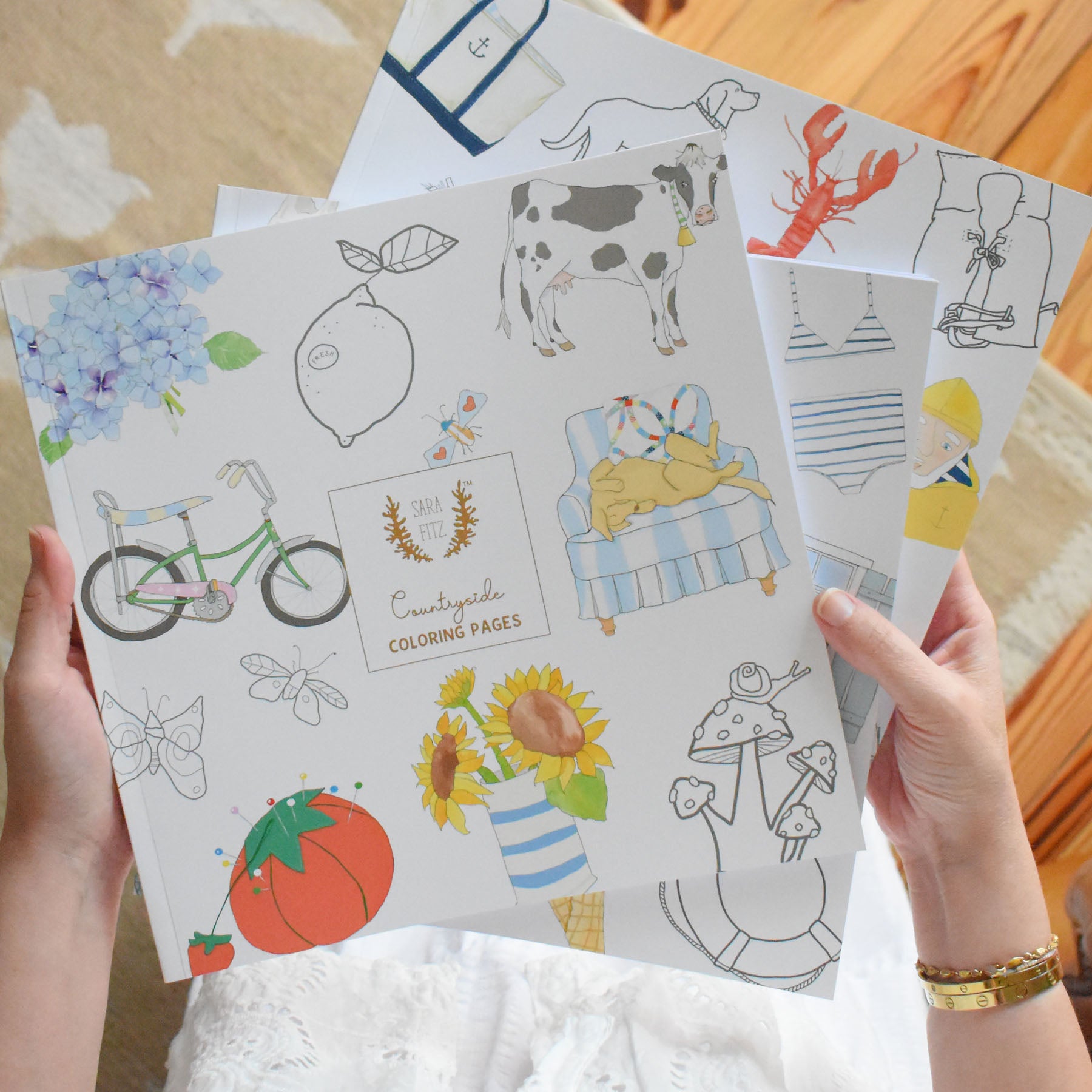 A person holds illustrated pages from the Sara Fitz Coloring Book Countryside, featuring charming New England-themed drawings—a bike, cow, lemon, chair, flowers, and insects—with additional adult coloring sheets from the book visible beneath them on a wooden floor.