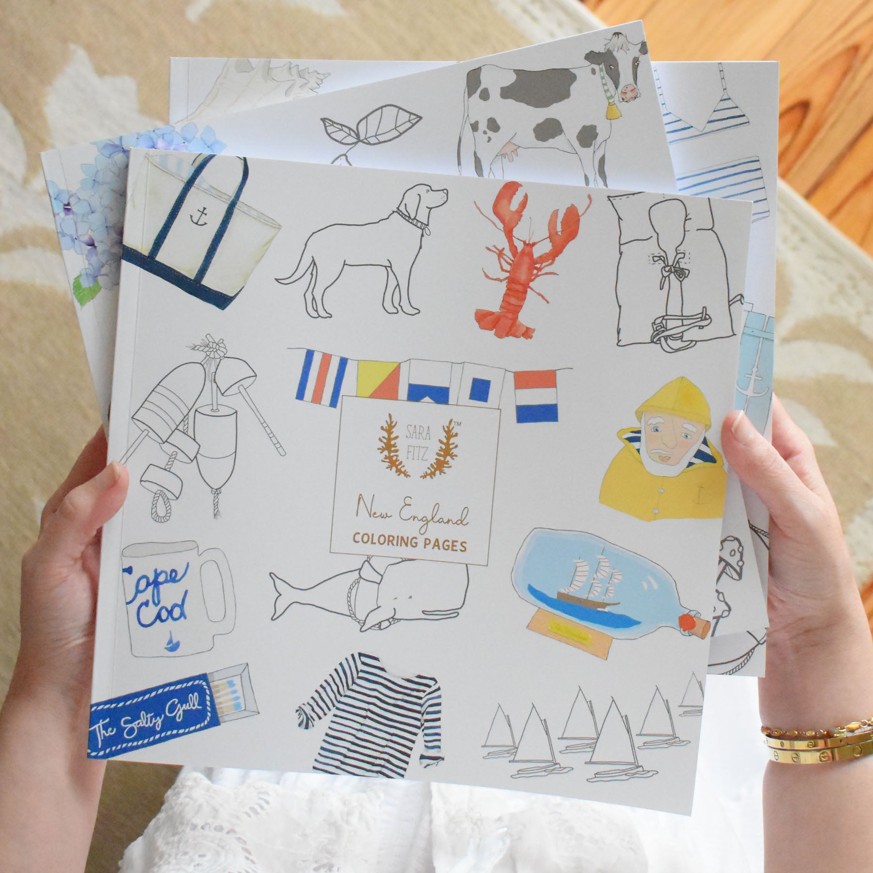 A person holds illustrated pages from the Sara Fitz “Coloring Book New England,” showcasing charming images such as a lobster, dog, sailor, ship in a bottle, Cape Cod sign, and nautical stripes. The bright and cheerful scene captures the classic New England spirit of this adult coloring book by Sara Fitz.
