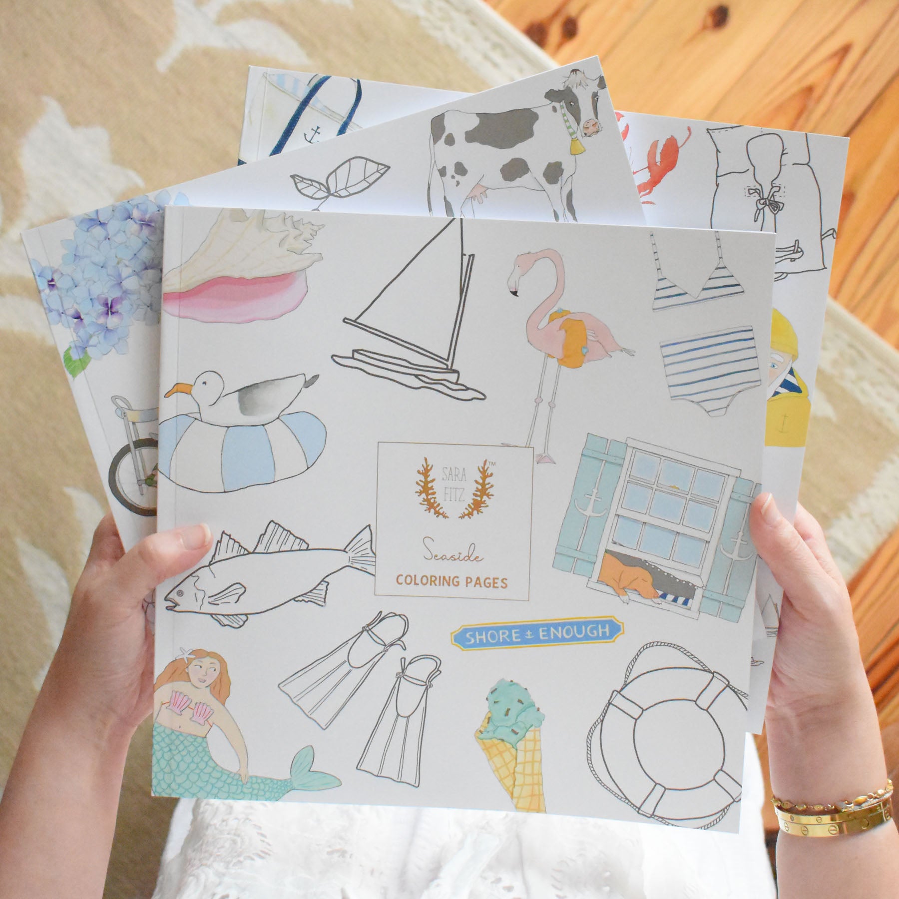 A person holds several illustrated Coloring Book Seaside editions by Sara Fitz, showcasing summer and seaside-themed New England illustrations—like sailboats, flamingos, swimming gear, animals, and ice cream—in the signature Sara Fitz style, all set against a wooden and carpeted background.