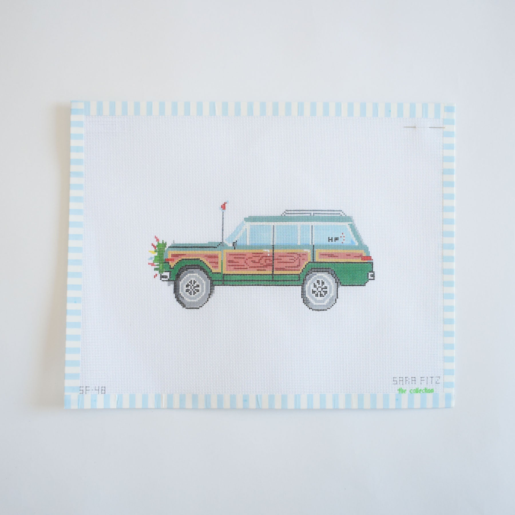 The Holiday Wagon Needlepoint by Sara Fitz showcases a vintage green station wagon with wood paneling, flowers in the back, and a flag on the antenna, bordered by blue and white stripes on 18 mesh canvas.