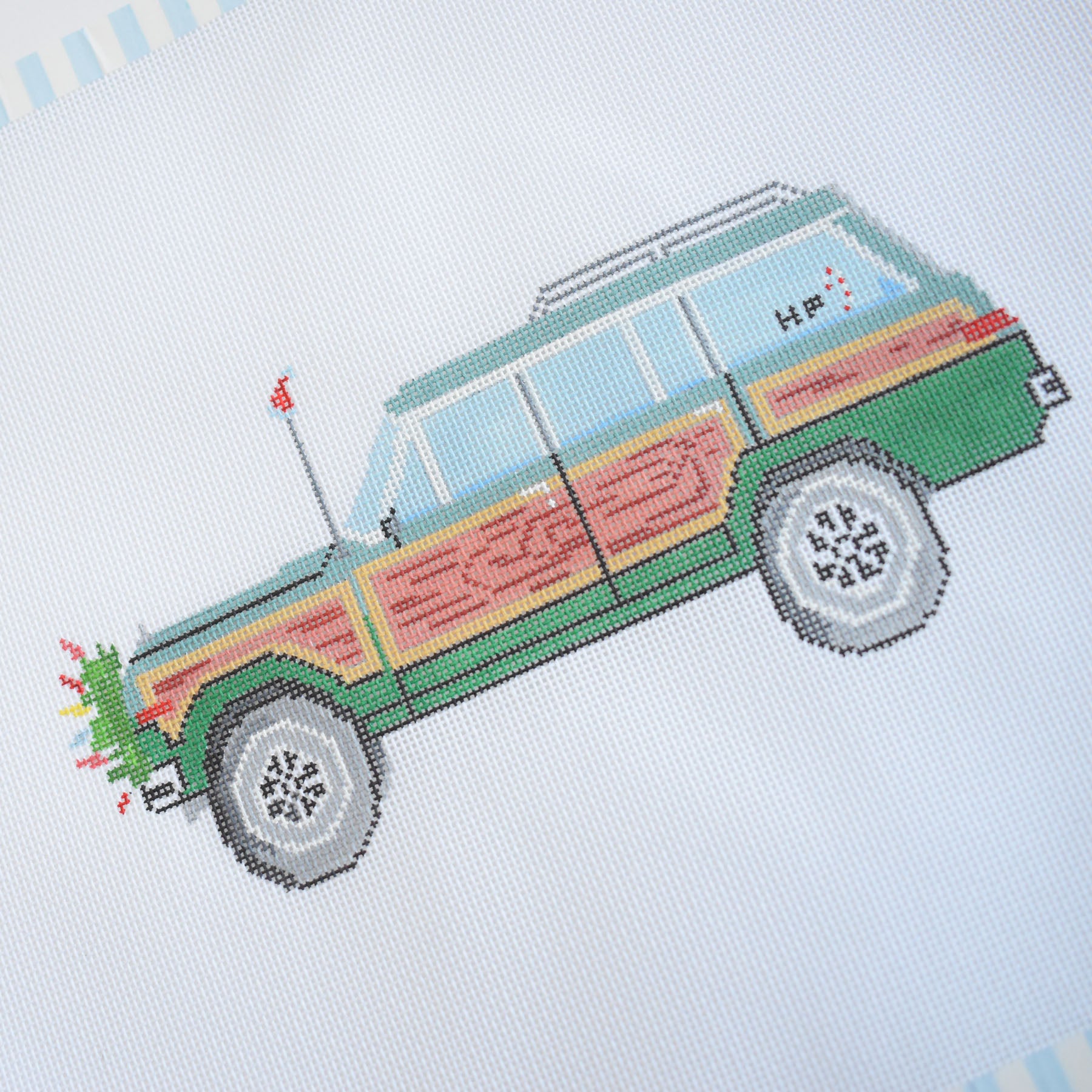 The Sara Fitz Holiday Wagon Needlepoint features a green station wagon with wood paneling, silver hubcaps, and a Christmas tree on top, designed on 18 mesh canvas with a blue and white striped border.