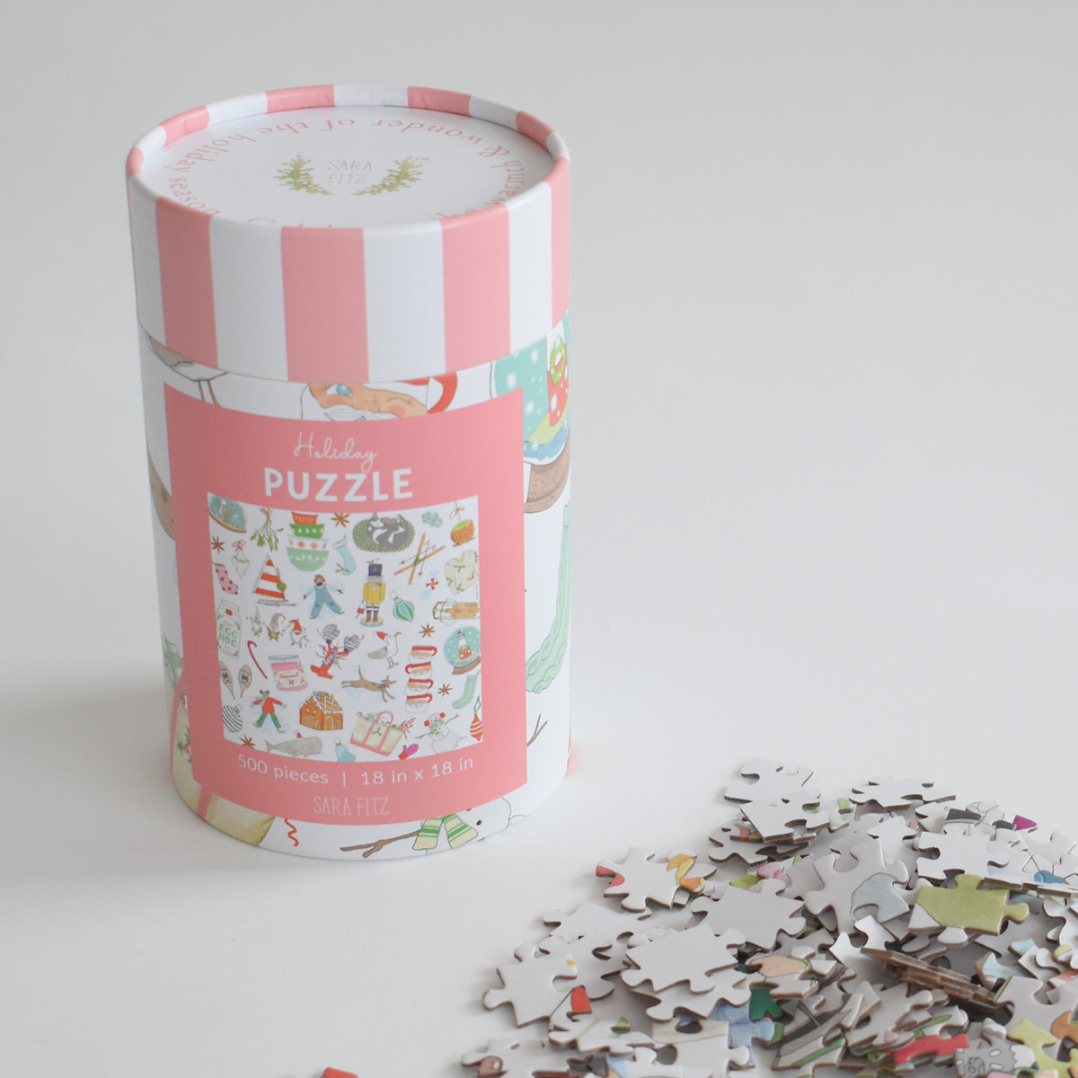 A cylindrical Sara Fitz Holiday Puzzle box with pink and white stripes, featuring an illustration of festive icons and a small pile of loose pieces beside it.