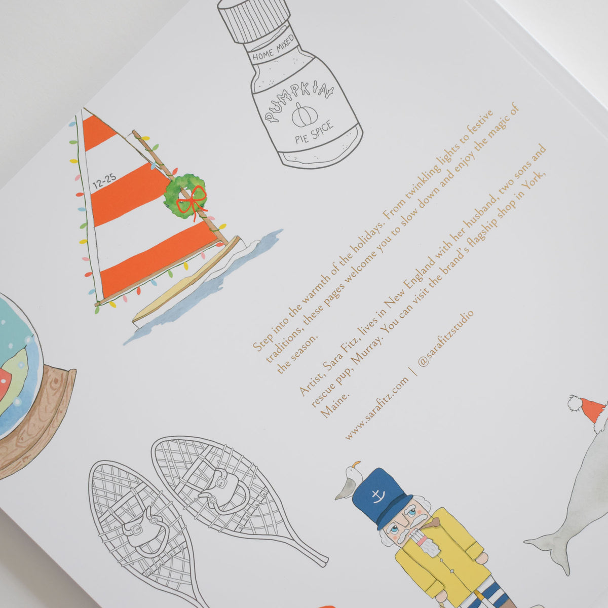A coastal coloring page from the Coloring Book Holiday by Sara Fitz featuring a sailboat with string lights, a jar of pumpkin pie spice, snowshoes, and a nutcracker, alongside descriptive text about the artist.