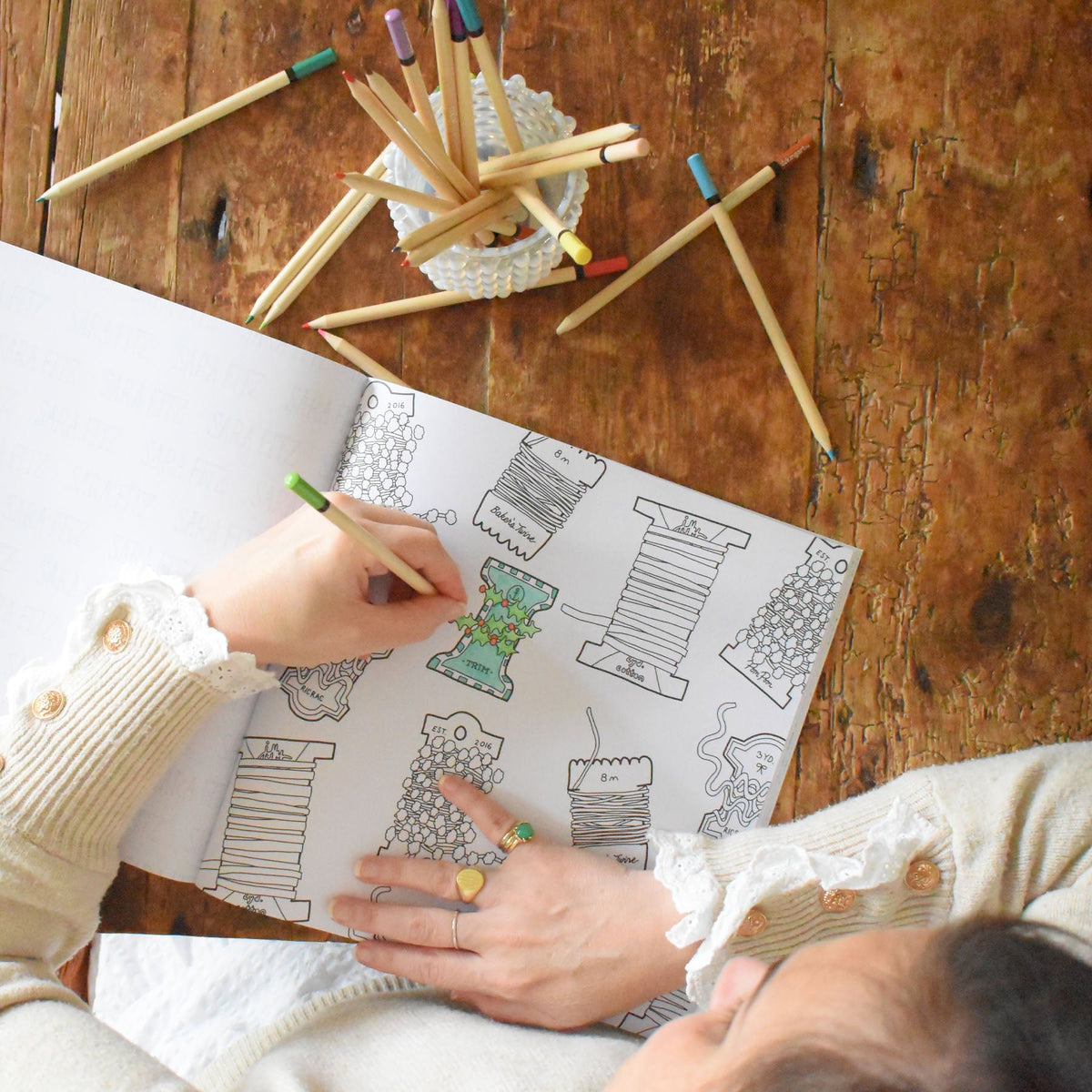 A person colors a page from a Sara Fitz Coloring Book Holiday featuring spool-themed coastal illustrations, with a glass of colored pencils nearby on a wooden table.