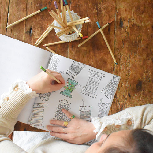 A person colors a page from a Sara Fitz Coloring Book Holiday featuring spool-themed coastal illustrations, with a glass of colored pencils nearby on a wooden table.