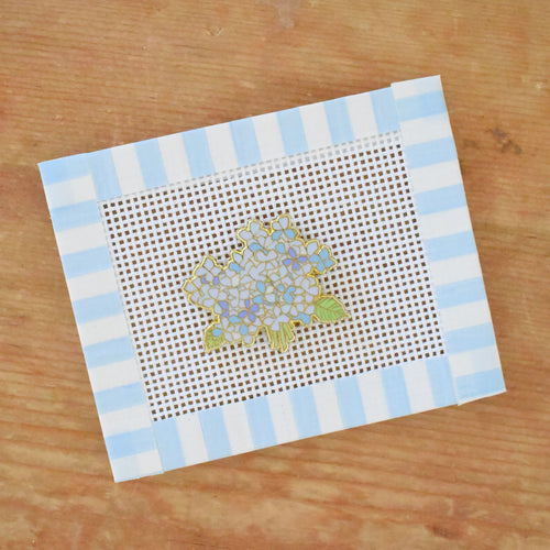The Sara Fitz Hydrangea Bloom Needle Minder is shown beside a hand-painted needlepoint canvas featuring a hydrangea flower with blue and white striped borders, all displayed on a wooden surface.