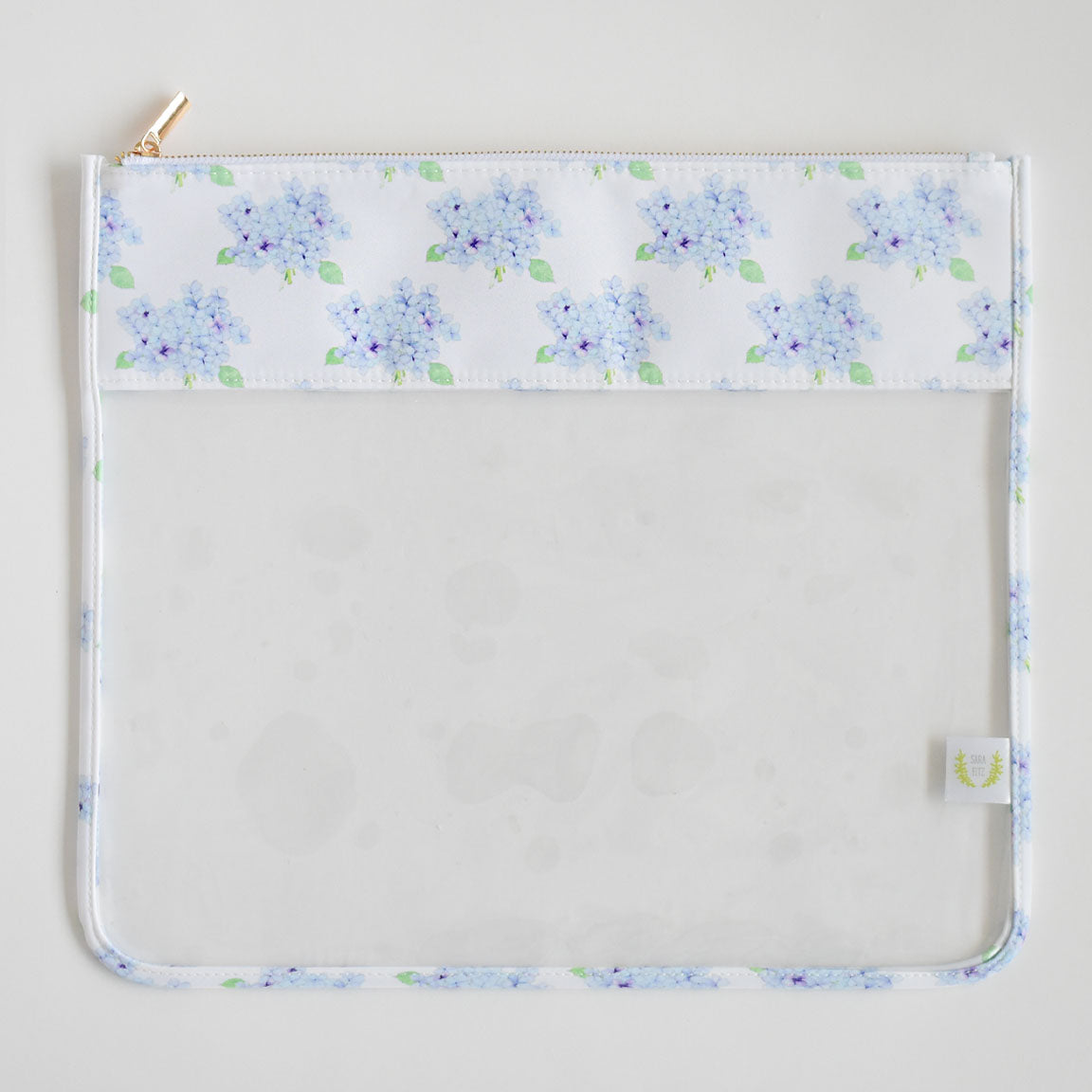 The Sara Fitz Hydrangea Project Bag is a clear plastic pouch with a white top border adorned with light blue hydrangea flowers and green leaves, featuring the charming Sara Fitz prints, a gold zipper closure, and a small fabric side tag.